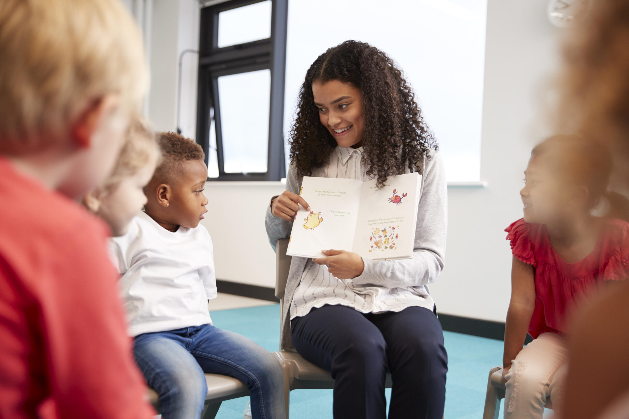 Young female teacher showing a picture in a book to kindergarten children sitting on chairs in a circle in the classroom, close up, selective focus
